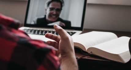 Fotografia di una scrivania con sopra un libro e un pc con una persona visibile nello schermo