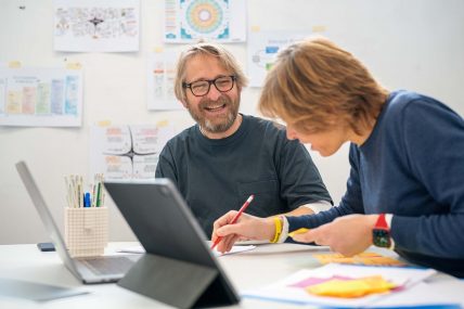Marco laughs and Ilaria writes on a sheet of paper in the office