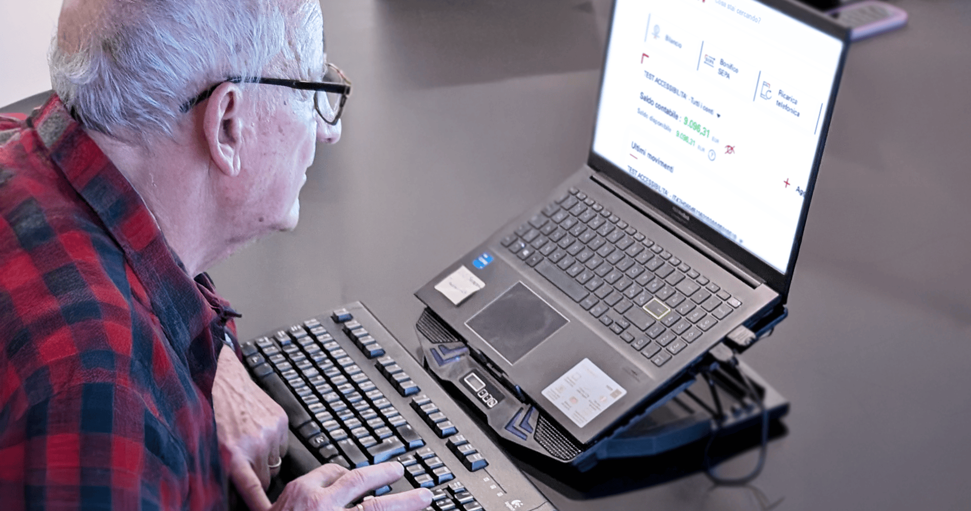 Uomo anziano usa tastiera esterna e schermo ingrandito per navigare nel sito della banca.